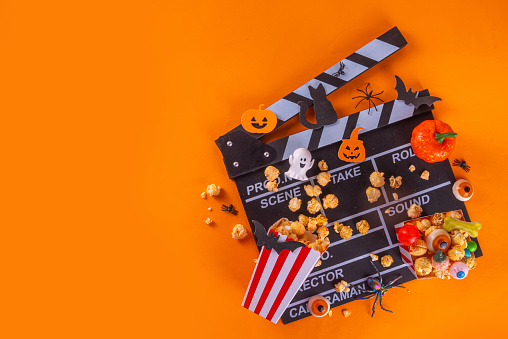 Horror movie, series night, Halloween cinema party with trick-or-treating sweets bucket, popcorn, movie clapperboard and Halloween decorations on high-colored background copy space