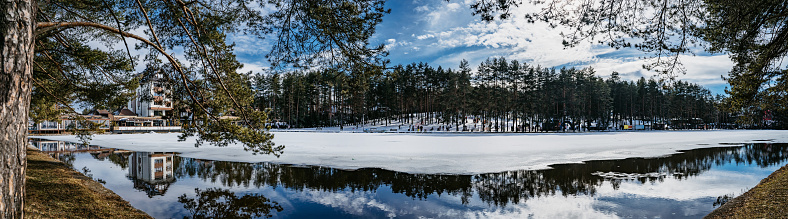 Winter landscape with beautiful reflection in the lake surrounded with trees and a ski resort during a sunny day.