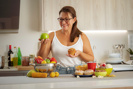 Woman holding an apple and a muffin making choice between junk food or healthy food