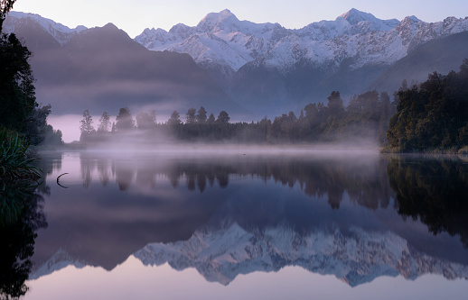 Dawn breaks over spectacular Lake Matheson on New Zealand's South Island. In the distance we see the snow capped peaks of the Southern Alps, reflected in the mirrorlike surface of the lake.