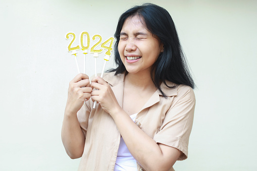 excited asian young woman celebrating new years eve by holding golden candles numbers 2024 with closed eye wearing casual brown shirt isolated on white background