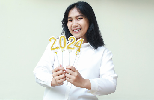 cheerful young female asian business woman smiling to camera by showing golden glittered 2024 candles for 2024 new years countdown celebration wearing formal white shirt isolated