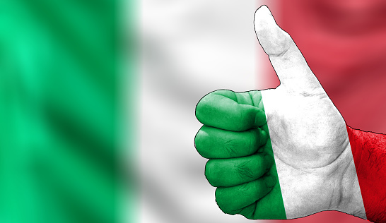 hand with thumbs up in approval with the Italian flag painted. Image with Italian flag background area out of focus, copy space area