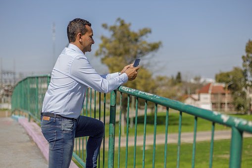 Latin man using his mobile phone leaning on the railing of a pedestrian bridge with copy space.