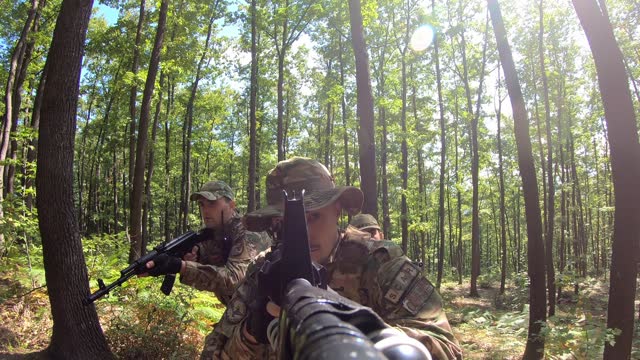 POV Close-up group of soldiers aiming with gun while moving through forest during military action. Squad of equipped fighters in camouflage uniform in forest.