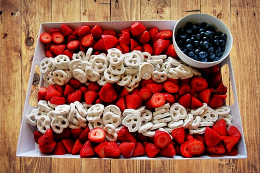 American fruit tray: fresh strawberries cut in half, a bowl of blueberries and Flipz pretzels with white syrup topping. On a wooden table, seen from above.