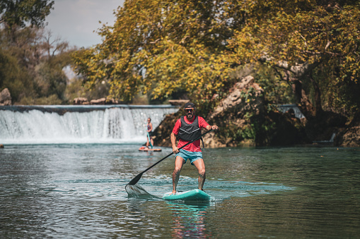 Mature couple floats on a SUP board along a forest river and waterfall. Summer active outdoor recreation.