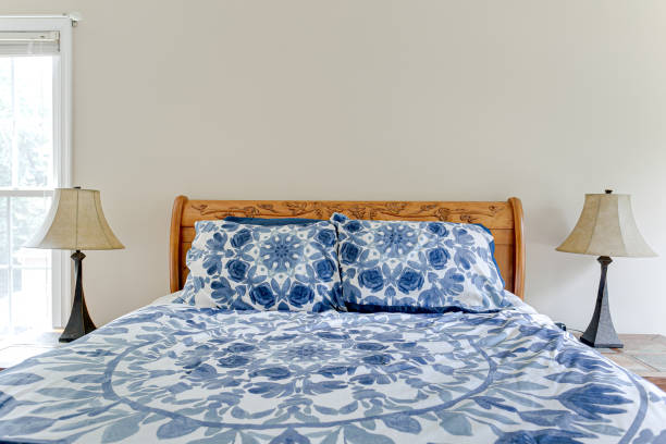 Serene bedroom with neatly made bed, blue and white comforter, floral pillows, lamp, and window exudes cozy tranquility.