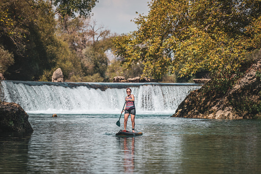 Active woman paddling SUP board on beautiful river by waterfall, autumn forest landscape and nature on background, stand up paddling water adventure outdoors, sport and healthy lifestyle concept