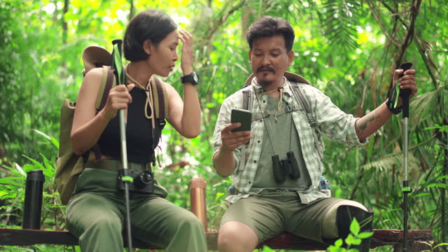Mid adult disabled man with prosthetic leg and binocular talking to Asian female while sitting on a tree trunk and discussion about direction planning on mobile phone for forest hiking, happy Asian couple spending the leisure time together