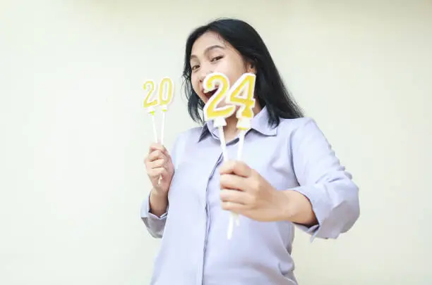 Photo of amazed young business woman asian screaming  with showing number 24 of 2024 number candle hold on hand, beautiful female wearing grey formal shirt, isolated, looking at camera, new year eve concept