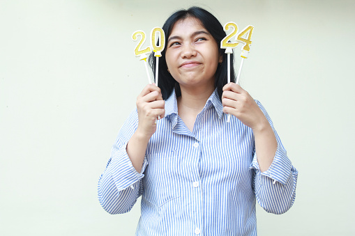 thoughtful asian young woman raising 2024 number candles holding on hand and looking above wearing blue stripes casual shirt, isolated on white, new years eve concept