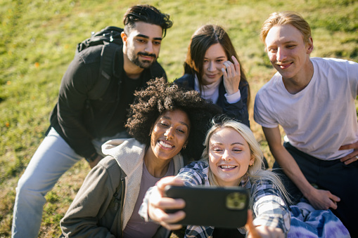 A college aged multiracial group take a self portrait together outside with a smartphone to capture the memory of a great day in a photo to post on social media.