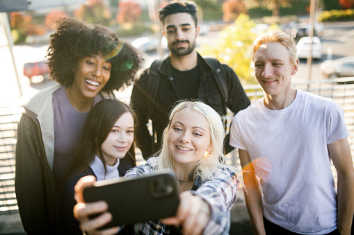 A college aged multiracial group take a self portrait together outside with a smartphone to capture the memory of a great day in a photo to post on social media.