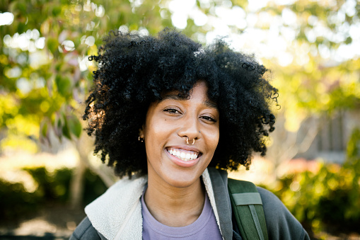 A portrait of a multiracial young woman at her school with a backpack full of textbooks and a big smile.  She sports a beautiful natural hair style.