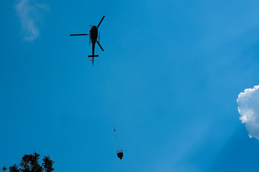 Helicopter with bag of water to put out fires flies over a forest area.