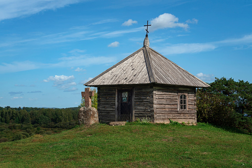 An old small wooden Orthodox chapel and a stone cross on Savkina Mountain on a sunny summer day, village Mikhailovskoye, Pushkinskie Gory, Pskov region, Russia