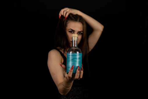 The black witch has prepared a magic potion for Halloween and is holding it out to the camera on a black background. A beautiful witch in the Gothic style holds a bottle of potion in her hand..