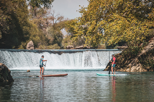 Two surfers walking on paddle boards in front of the waterfall