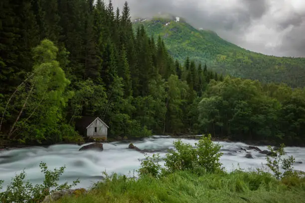 Rapids at a mountain creek flow along the Norwegian scenic route Gaularfjellet between Moskog and Balestrand during an overcast, stormy day. A lone wood house stands near the edge.