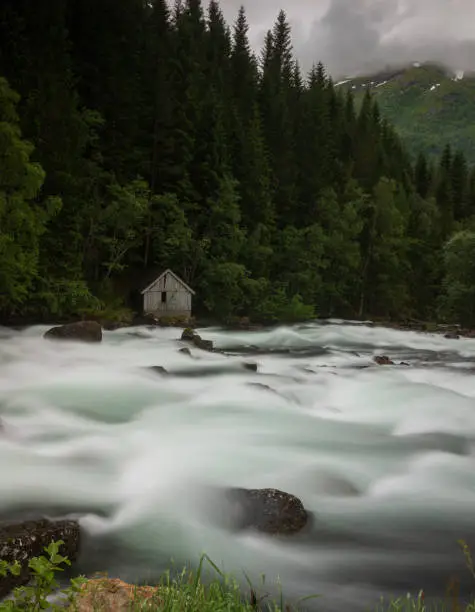 Rapids at a mountain creek flow along the Norwegian scenic route Gaularfjellet between Moskog and Balestrand during an overcast, stormy day. A lone wood house stands near the edge.