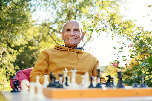 Portrait of smiling positive older man seated at chessboard in cottage garden on nature background