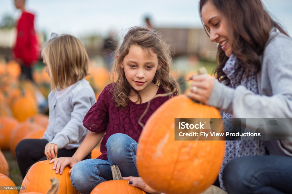 Mom and young daughters examine pumpkins at a farm Beautiful young mother sits with her elementary school age daughter's in a pumpkin patch, examining a large pumpkin while spending the day together on a beautiful October day. Day Stock Photo