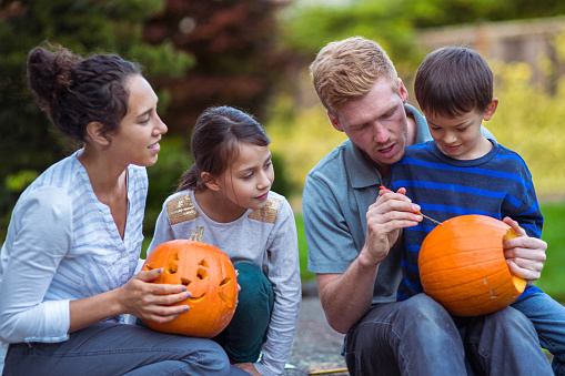 Multiracial family of four sit on the driveway of their home, carving pumpkins together as they enjoy a warm autumn day. The young boy sits on his father's lap, watching him carve.