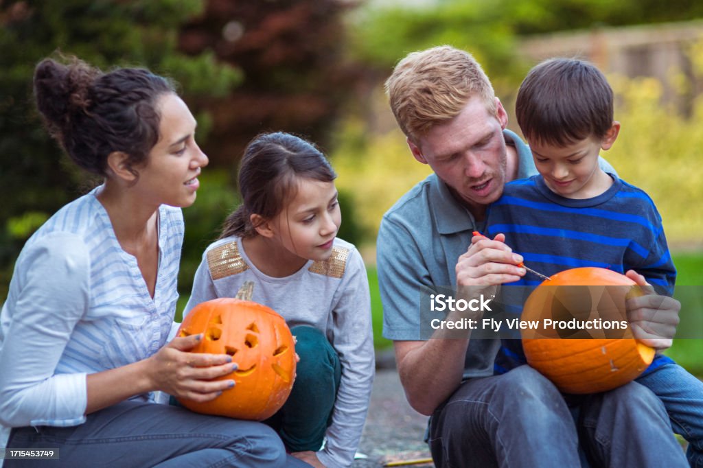 Family of four carve pumpkins outside their home Multiracial family of four sit on the driveway of their home, carving pumpkins together as they enjoy a warm autumn day. The young boy sits on his father's lap, watching him carve. Choosing Stock Photo