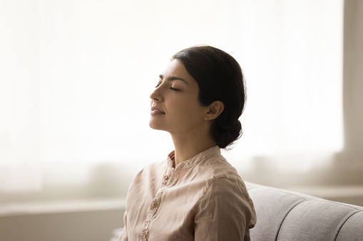 Calm tranquil young Indian woman meditating at home, sitting on sofa with straight back and closed eyes, breathing fresh air, doing yoga breathwork, practicing relaxation, stress relief, meditation