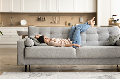 Peaceful relaxed young Indian woman resting on grey soft couch with closed eyes on modern studio apartment interior, breathing, sleeping, enjoying relaxation, fresh cool air, comfort