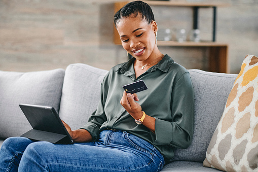 Woman holding credit card and shopping online with digital tablet on sofa at home, smiling and looking at card