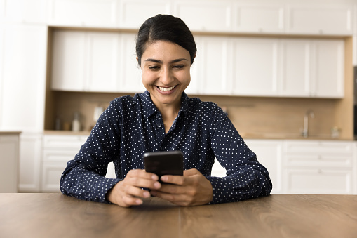 Cheerful beautiful Indian business woman using online application on mobile phone, having fun, laughing, typing on digital gadget, making video call, enjoying internet communication