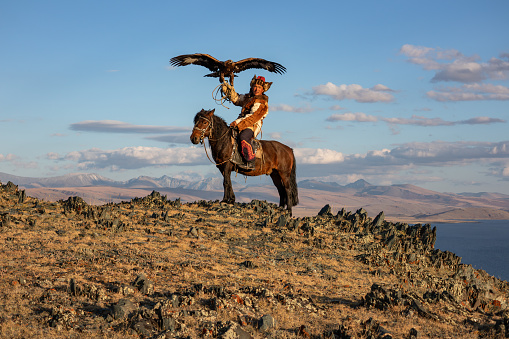 A Kazak eagle hunter sitting on his horse in the Altai Mountains, in evening sunlight. The hunter's large golden eagle is held on his arm, with it's wings open. The eagle hunter is wearing traditional Kazak clothing and a fur lined hat.