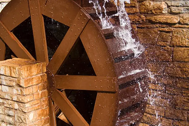 A wooden water wheel or watermill turbine milling, grinding, turning, and generating power, perhaps as a traditional alternative energy source. Positioned against the stone wall of a cottage building exterior, located in St. Paul, Minnesota, USA.