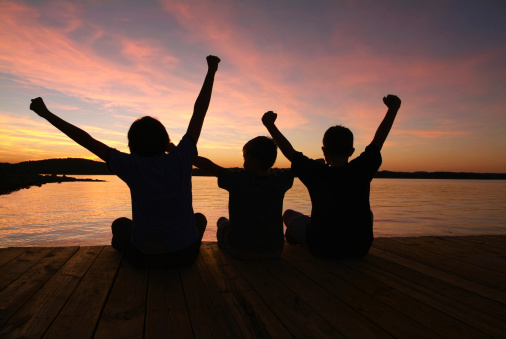 Three happy young children raising their arms in celebration. They are sitting on a wooden dock that is floating on a lake. The sun is setting in front of them creating brilliant colors in the sky and reflecting onto the water. The children are in silhouette. 