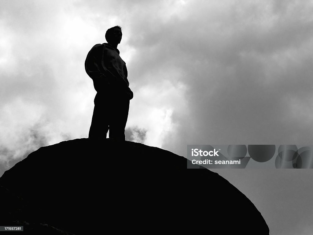 Top of the World 2 A shot of a young man standing on top of a large rock and surveying the land.  The figure is unrecognizeable and mostly in silhouette against the cloudy sky.  Photo is in black and white. In Silhouette Stock Photo