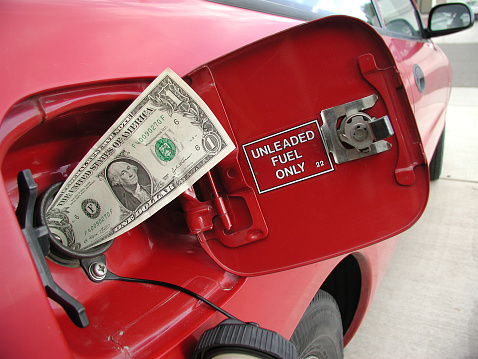 This landscape-orientation color photo shows a concept on rising fuel prices or gas prices. The image has a one-dollar bill sticking out of a sporty red car’s gas tank, showing the George-Washington side of the money. The inside of the gas hatch says “unleaded fuel only.”