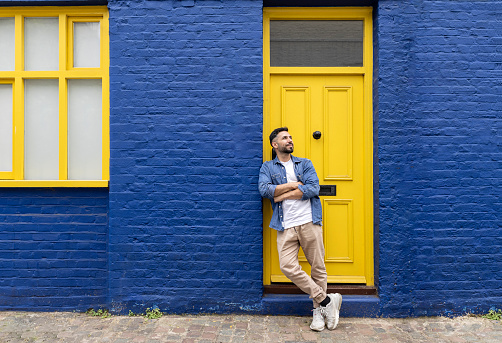 young Latino adult waits with crossed arms for his appointment leaning on a blue wall with a yellow door typical of London.