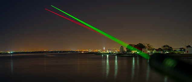 2 visible beams of laser light, red and green, focusing in the direction of Lisbon, crossing the Tagus River from the city of Barreiro.  night image.