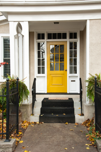 A yellow door on the entrance to a residential home in the summer with potted ferns on the side of the steps to the front door.