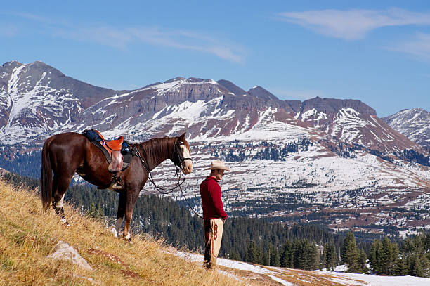 a man wearing a red shirt stands in profile looking at view with his horse in a mountain meadow with rugged mountainside ridges and peaks on the horizon underneath a cloud filled blue sky.  such beautiful nature scenery and outdoor recreation and adventure can be found in the san juan range of the colorado rocky mountains.  horizontal composition taken in durango, colorado.