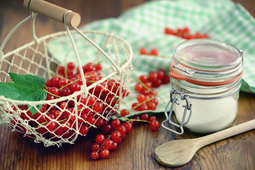 white metal basket with lot red currant and a glass with sugar on table with checked dish towel, ready to be prepared to make jam, home botting. kitchen utensil like wooden spoon. cross-processed. 