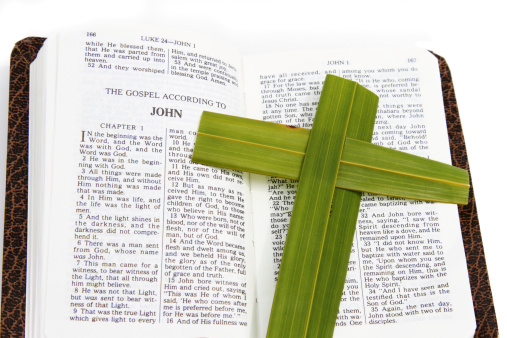 A New Testament Bible is open to the Gospel according to John. A cross made from palm leaves lies on one side of the open book.