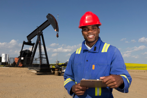 A royalty free image from the oil industry of a friendly oil worker, holding a clipboard, in front of a pumpjack.