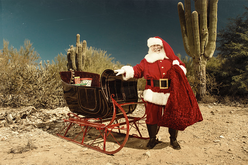 Santa Claus standing next to his sleigh in a desert setting surrounded by cacti.  Santa Claus has one hand on a sleigh and is looking forward.  The ground is sandy and along with the cacti desert brush and bushes can be seen.