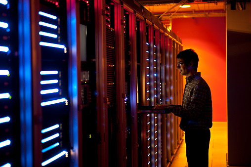 The interior of a modern server room in a data center.  An IT engineer is busy configuring the servers. The room is dark, but the servers themselves are lit.  The servers at the left side are lit in blue, while the ones at the other end are lit by an orange light source coming from the right side.