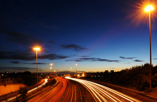 Twilight image of motorway rush hour traffic with trails of light caused by the long exposure. Overhead at top of frame are a series of dots, which are the wing lights of a passenger flight coming into land at a nearby airport.