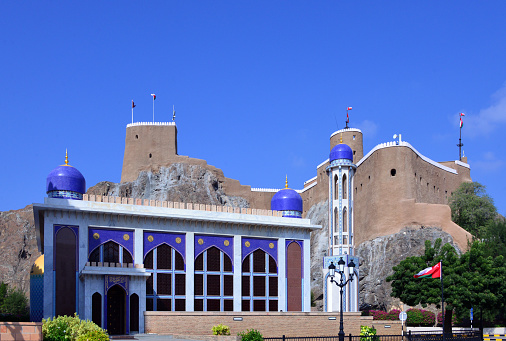 Old Muscat, Oman: Al Khor Mosque with its blue domes on Bab al Muthaib Street. Although successive renovations made it look modern, the mosque is hundreds of years old, it was once known as Masjid Al-Shuhadaa, or the Mosque of Martyrs, for the Omanis who died in battle against the Portuguese. Senior members of the Omani royal family, including the Sultan pray here. In the background the towers and walls of Al-Mirani Fort, a Portuguese 16th century fortress.