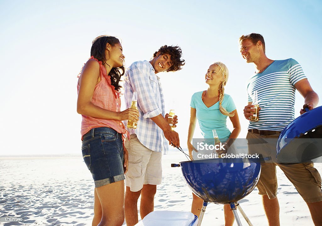 Enjoying some good food and fresh air Four young people standing around a barbeque on the beach Adult Stock Photo